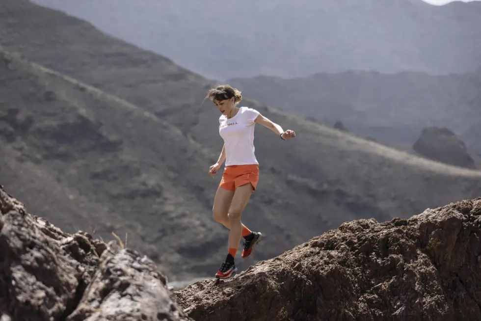 Denisa Dragomir Merrell runner with a white shirt and orange shorts running on rocks